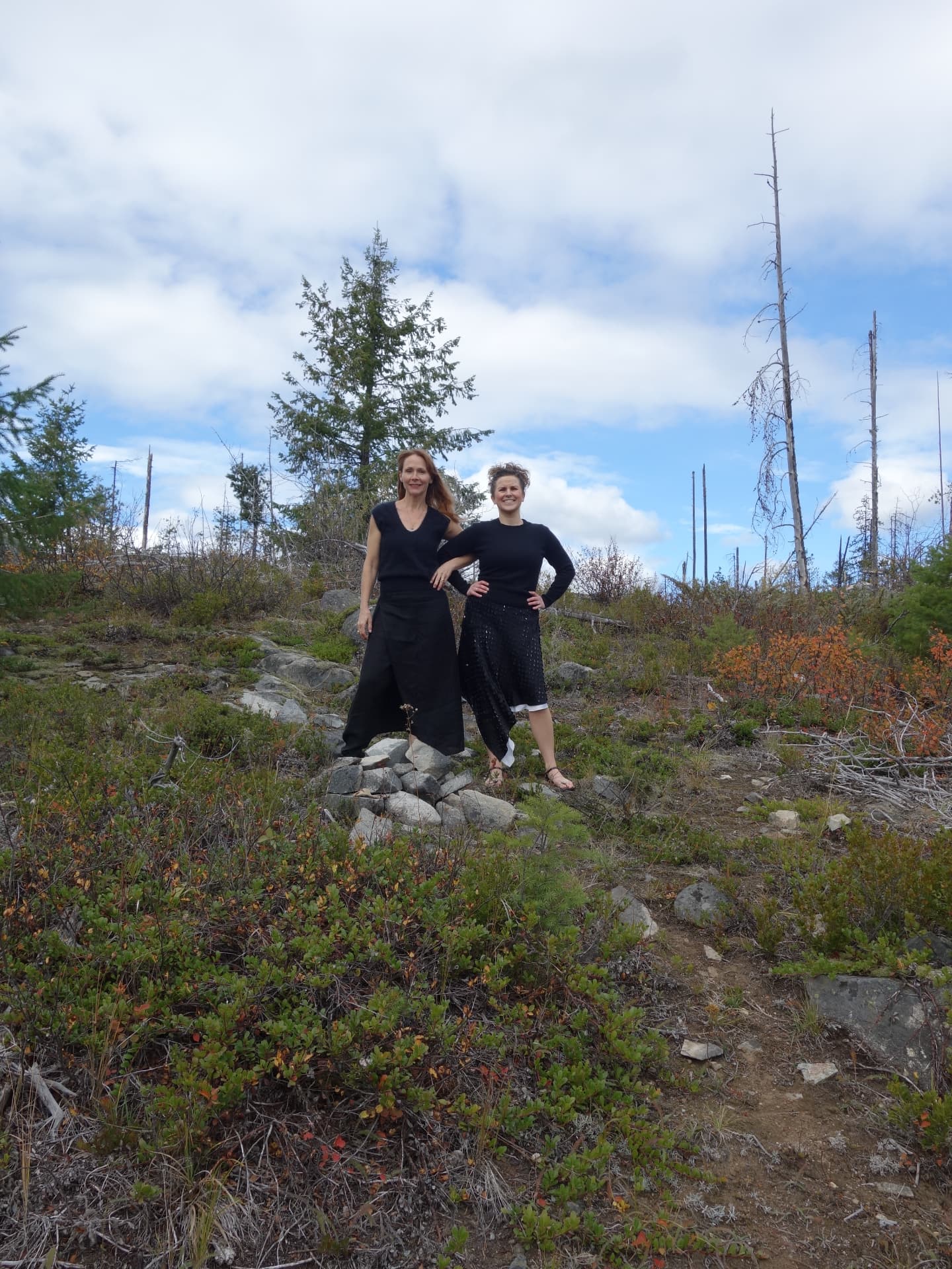 Two women standing together on a rocky hilltop among low shrubs and bare trees