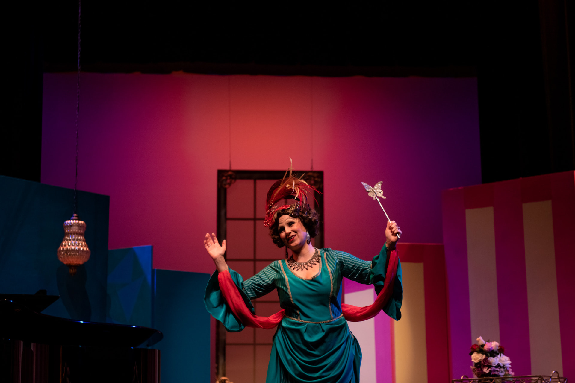 Performer in a teal gown and feathered headpiece raising a wand