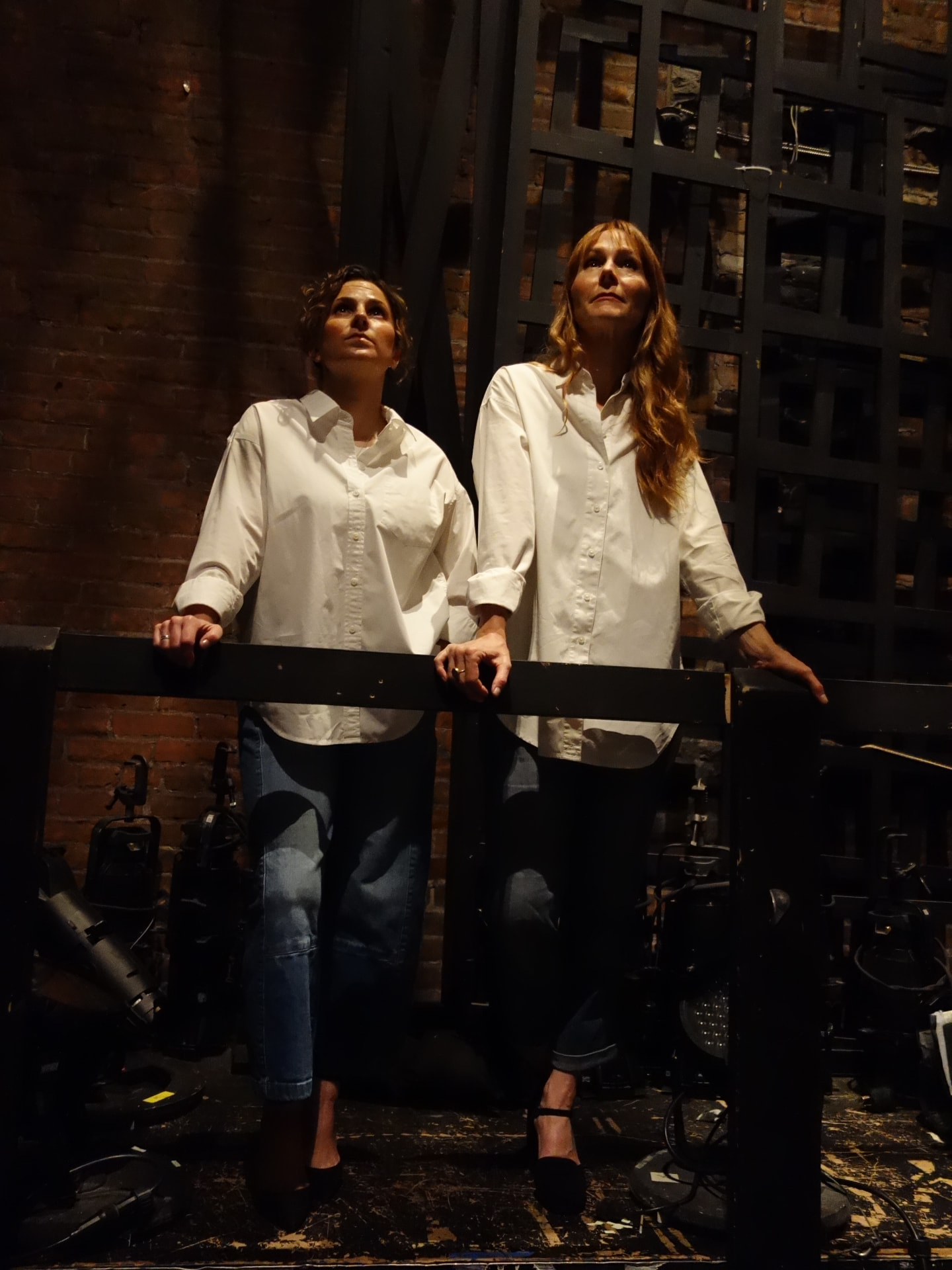 Lindsay Clague and Rachel DeShon standing together at a railing on a theatre stage, looking up toward the lights