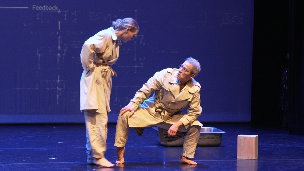 Two actors in trench coats on a dark stage